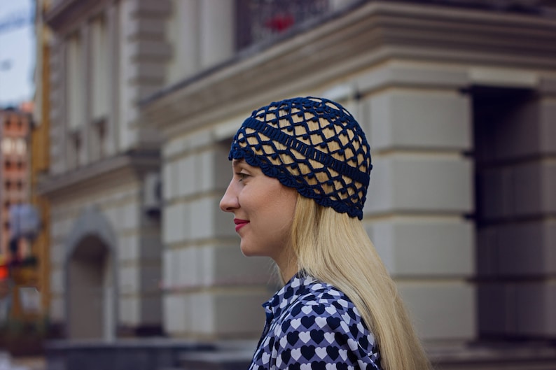 May include: A woman wearing a blue crocheted hat with a geometric pattern. The hat is worn on her head and covers her ears. She has long blonde hair and is wearing a black and white patterned shirt.