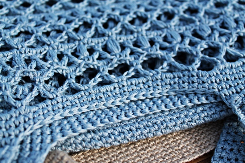May include: A close-up of a blue crocheted fabric with a textured pattern. The fabric is folded over itself, revealing a layer of beige fabric underneath.
