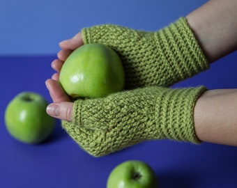 May include: A pair of green crocheted fingerless gloves with a green apple in the palm of one hand. Three green apples are on a purple surface.