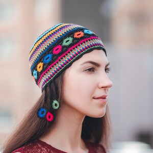 May include: A woman wearing a colorful crocheted hat with black tassels. The hat has a geometric pattern with circles and stripes in shades of pink, blue, yellow, and green.