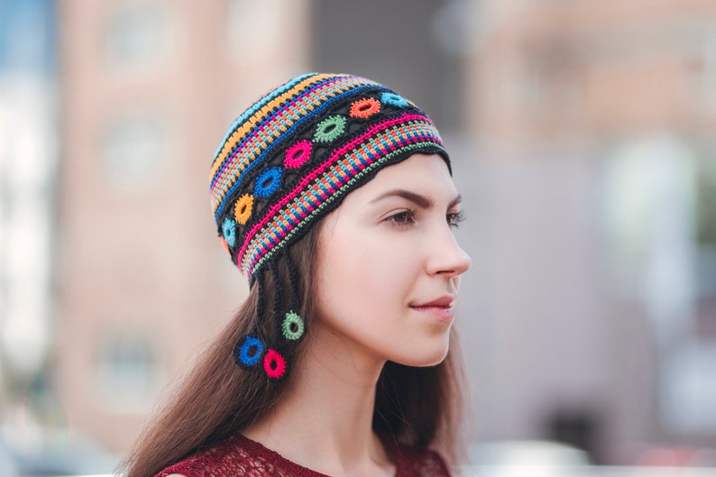 May include: A woman wearing a colorful crocheted hat with black tassels. The hat has a geometric pattern with circles and stripes in shades of pink, blue, yellow, and green.