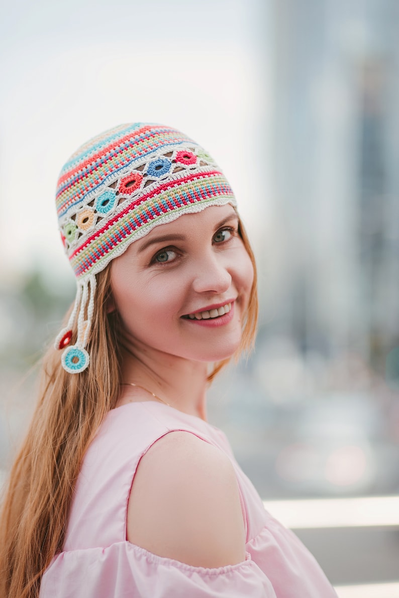 May include: A woman with long, light brown hair is wearing a pink, off-the-shoulder top and a colorful crocheted hat with a white base and colorful stripes and circles.
