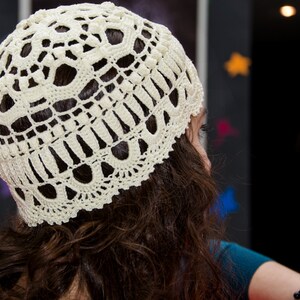 May include: A white crocheted hat with a delicate lace pattern. The hat is worn by a person with long brown hair.
