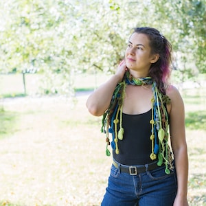 May include: A woman wearing a black tank top and blue jeans with a colorful crocheted scarf with dangling beads and leaves.