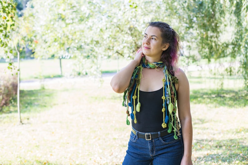 May include: A woman wearing a black tank top and blue jeans with a colorful crocheted scarf with dangling beads and leaves.