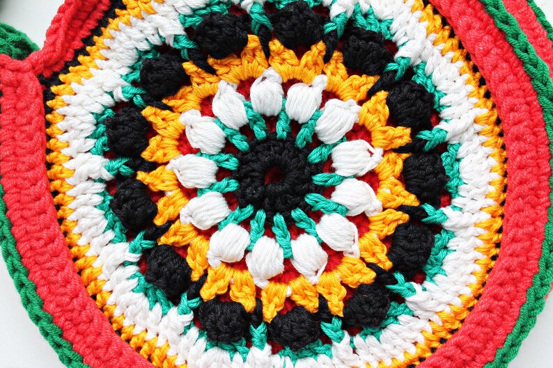 May include: Close-up of a colorful crocheted round mat with a central design of black, white, green, and yellow yarn. The mat has a red border with a green and white trim.