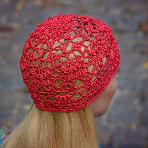 May include: A red crocheted hat with a lacy design. The hat is worn on a person's head, with their blonde hair visible.