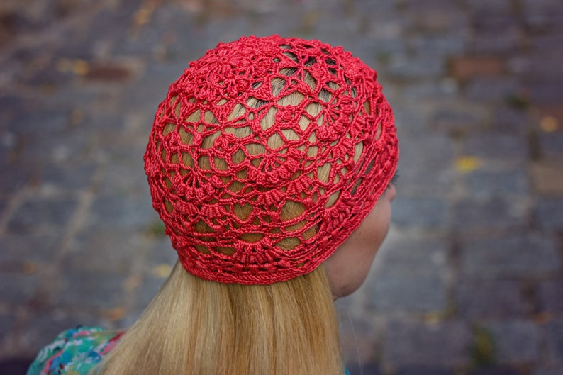 May include: A red crocheted hat with a lacy design. The hat is worn on a person's head, with their blonde hair visible.