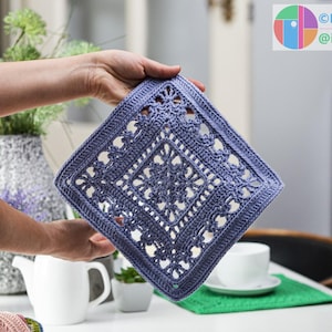 May include: A square, crocheted doily in a dusty blue hue, featuring an intricate openwork design. The doily is held up, showcasing its detailed floral and geometric patterns. The image includes a white teacup and saucer, and a green mat.