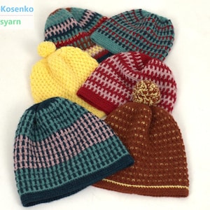 Op de afbeelding: Een verzameling handgebreide mutsen in verschillende kleuren, waaronder geel, blauwgroen, rood en bruin. Elke hoed heeft een textuurpatroon en sommige zijn versierd met pompons. De hoeden zijn gerangschikt op een witte ondergrond.