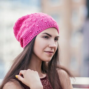 A woman stands partially turned, displaying an exquisite handmade crochet hat with an intricate pattern.