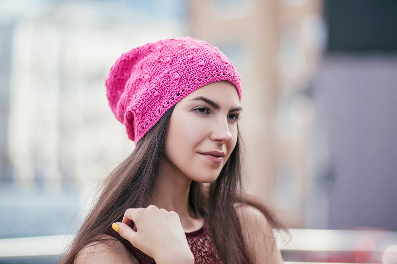 A woman stands partially turned, displaying an exquisite handmade crochet hat with an intricate pattern.