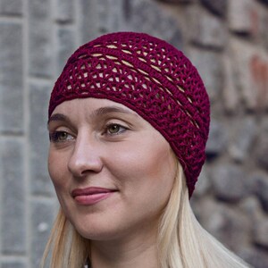 May include: A close-up of a woman wearing a burgundy crocheted beanie hat. The hat has a lacy design and is made with a soft yarn.