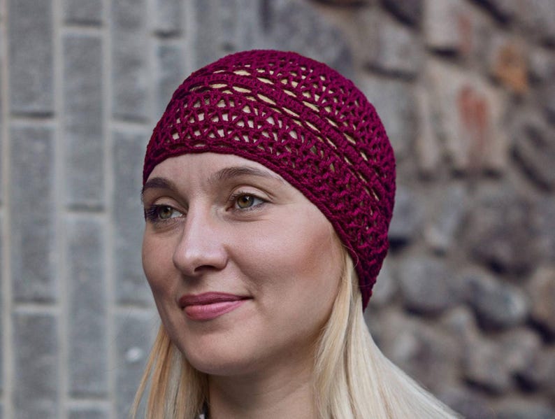 May include: A close-up of a woman wearing a burgundy crocheted beanie hat. The hat has a lacy design and is made with a soft yarn.