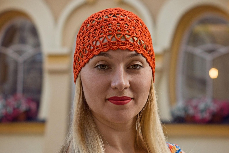 May include: A woman with long blonde hair is wearing a bright orange crocheted hat. The hat has a lacy design and is worn on her head.