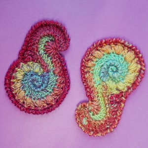 Könnte beinhalten: Zwei handgefertigte Häkelapplikationen in Paisley-Form. Die Applikationen weisen ein Spiraldesign mit einer Mischung aus Farben auf, darunter Rot, Gelb, Grün und Blau. Die Ränder sind dunkler rot und der Hintergrund ist einfarbig lila.