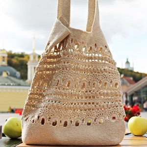 May include: A beige crocheted tote bag with a large open weave pattern. The bag has two long straps and is sitting on a wooden table.