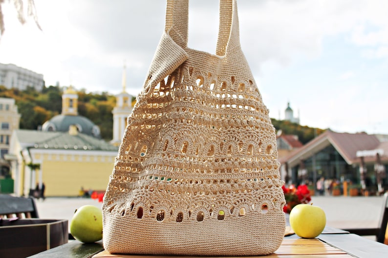 May include: A beige crocheted tote bag with a large open weave pattern. The bag has two long straps and is sitting on a wooden table.