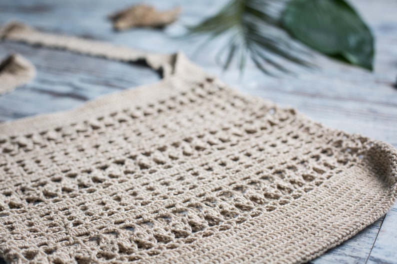 May include: A close-up of a beige crocheted tote bag with a simple, open-weave design. The bag is made of natural fibers and has a relaxed, bohemian style.