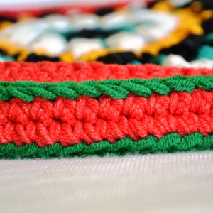 May include: Close-up of a red and green crocheted border. The border is made of yarn and has a textured, woven look.