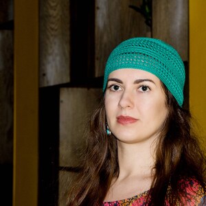 May include: A woman with long brown hair wearing a teal crocheted beanie hat. She is looking at the camera with a neutral expression.