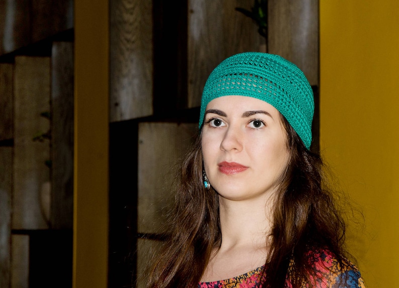 May include: A woman with long brown hair wearing a teal crocheted beanie hat. She is looking at the camera with a neutral expression.