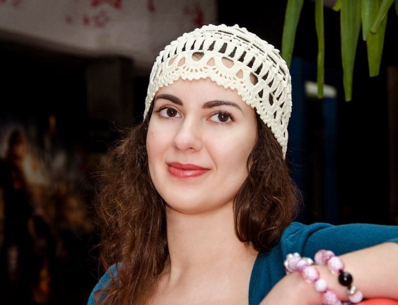 May include: A woman with long brown hair wearing a white crocheted hat. She is smiling and looking at the camera. She is wearing a blue shirt and has bracelets on her wrist.