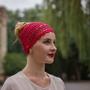 May include: A woman with red lipstick is wearing a red crocheted headband. She is looking to the right and has her hair pulled back.