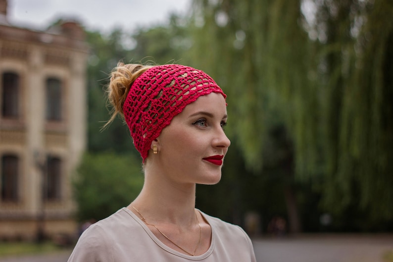 May include: A woman with red lipstick is wearing a red crocheted headband. She is looking to the right and has her hair pulled back.