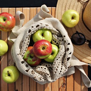 May include: A crocheted mesh bag filled with green and red apples. The bag is on a wooden surface next to a straw hat and sunglasses.