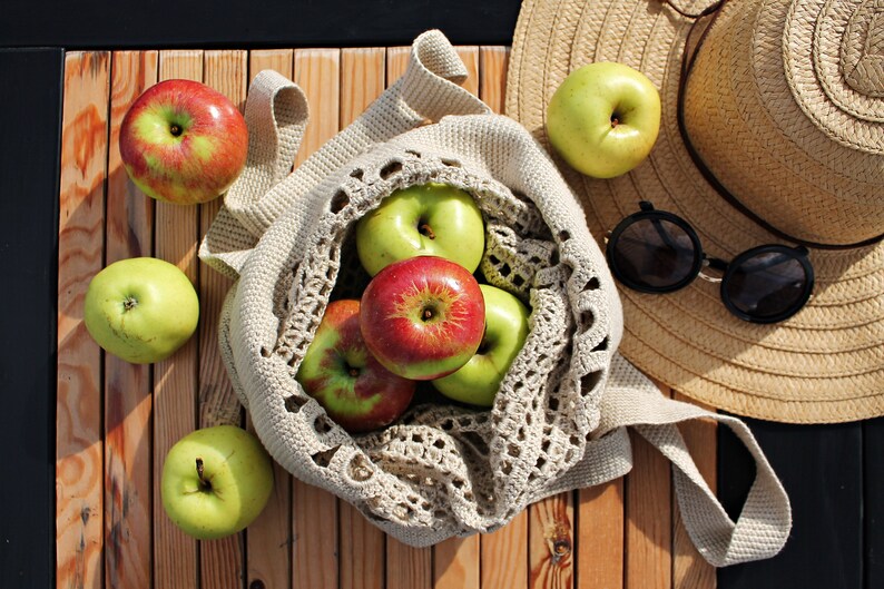 May include: A crocheted mesh bag filled with green and red apples. The bag is on a wooden surface next to a straw hat and sunglasses.