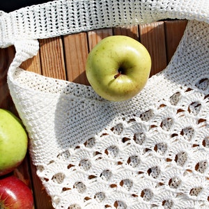 May include: A white crocheted tote bag with a lacy design, filled with green apples. The bag is lying on a wooden surface.