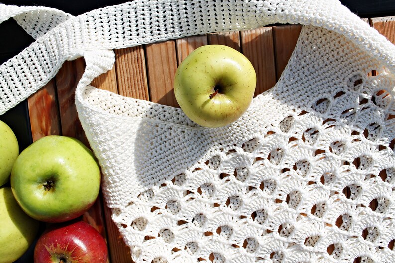 May include: A white crocheted tote bag with a lacy design, filled with green apples. The bag is lying on a wooden surface.
