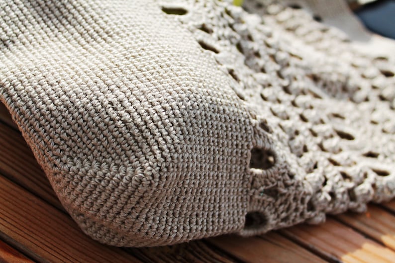 May include: Close-up of a beige crocheted bag with a textured pattern. The bag has a square shape and a crocheted design with openwork details.
