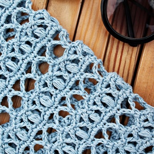 May include: A close-up of a light blue crocheted fabric with a delicate pattern. The fabric is laid out on a wooden surface.