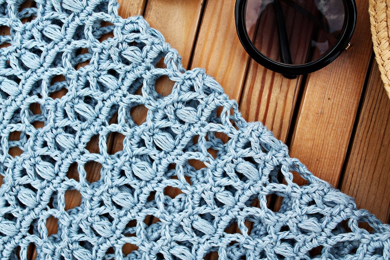 May include: A close-up of a light blue crocheted fabric with a delicate pattern. The fabric is laid out on a wooden surface.