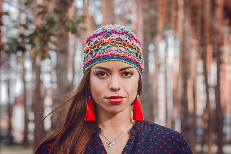 May include: A woman wearing a colorful crocheted hat with red tassel earrings. She is wearing a black and white polka dot shirt.