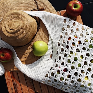 May include: A white crocheted tote bag with a straw hat and apples. The bag has a large open weave pattern.