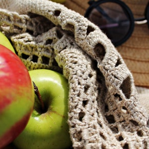 May include: A close-up of a crocheted bag filled with red and green apples. The bag is made of a beige crochet fabric with a delicate pattern. The apples are arranged in a pile, with the red apple on top.