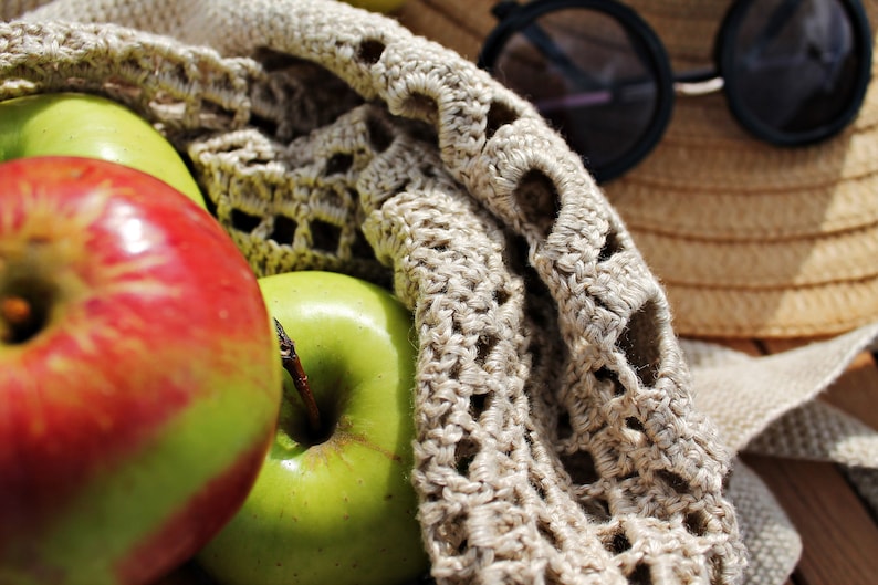 May include: A close-up of a crocheted bag filled with red and green apples. The bag is made of a beige crochet fabric with a delicate pattern. The apples are arranged in a pile, with the red apple on top.