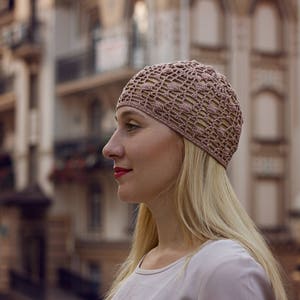 May include: A woman with long blonde hair is wearing a light pink crocheted hat. She is looking to the right and has a neutral expression on her face.