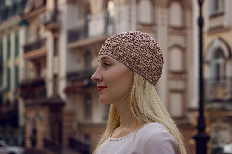 May include: A woman with long blonde hair is wearing a light pink crocheted hat. She is looking to the right and has a neutral expression on her face.