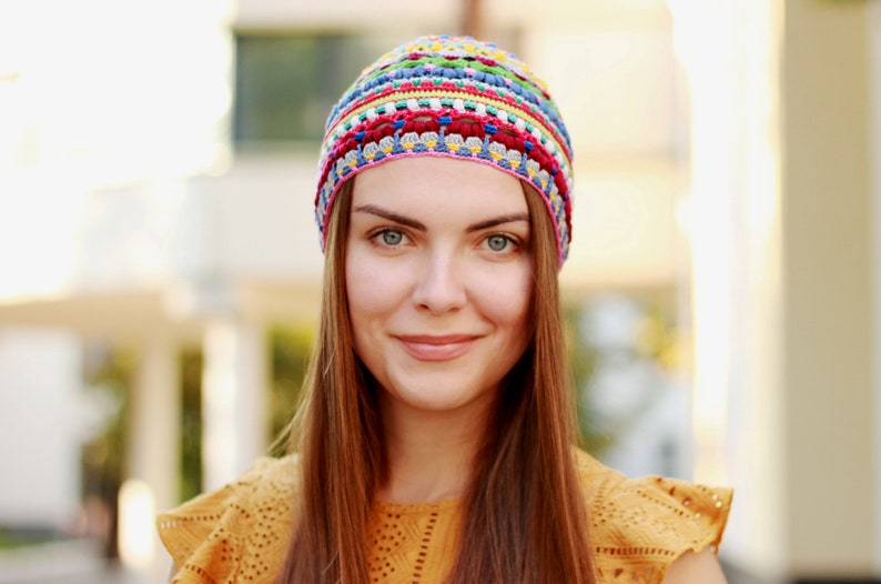 The woman wears a crocheted hat made from fine yarn and smiles against the backdrop of an urban landscape.
