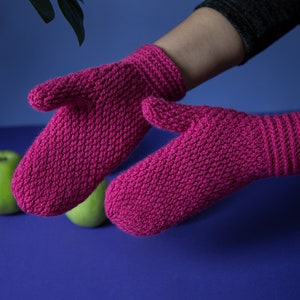 Könnte beinhalten: Ein Paar rosa gestrickte Fäustlinge mit einem strukturierten Muster. Die Fäustlinge werden vor einem blauen Hintergrund gezeigt.