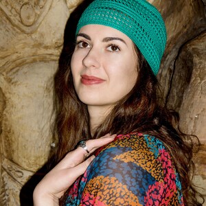 May include: A woman with long brown hair wearing a teal crocheted hat and a colorful patterned shirt with a leopard print design. She is wearing several bracelets on her wrist.