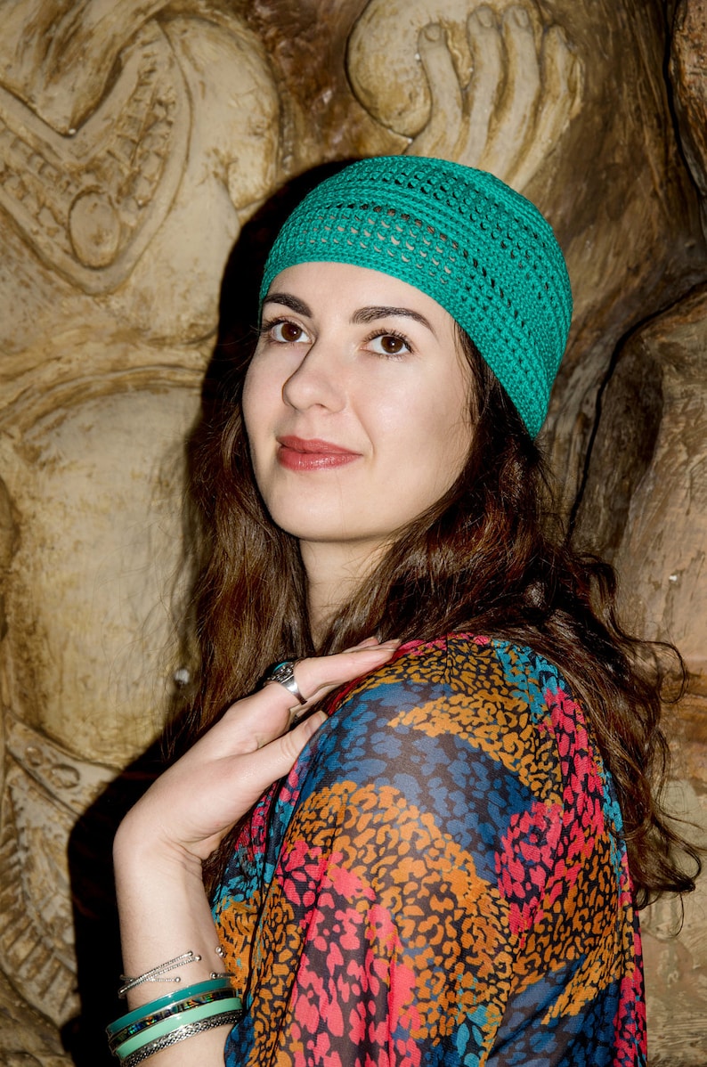 May include: A woman with long brown hair wearing a teal crocheted hat and a colorful patterned shirt with a leopard print design. She is wearing several bracelets on her wrist.
