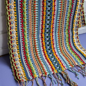 A very beautiful striped crochet scarf pattern with tassels is showcased on light purple background