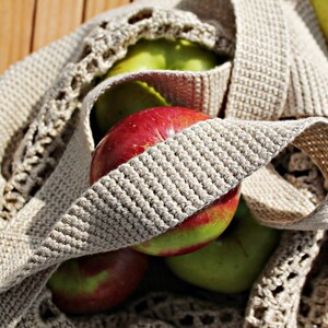 May include: A close-up of a crocheted bag filled with red and green apples. The bag is made of a beige crocheted material with a textured pattern. The apples are arranged in a cluster, with some visible through the open weave of the bag.