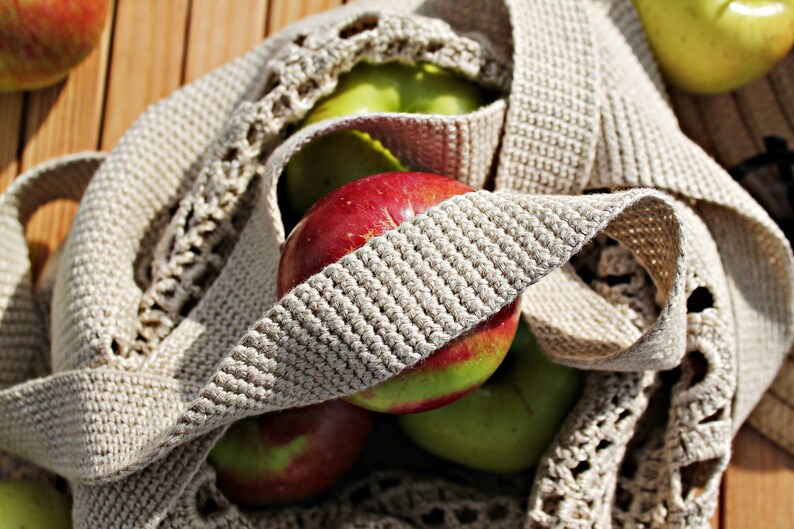 May include: A close-up of a crocheted bag filled with red and green apples. The bag is made of a beige crocheted material with a textured pattern. The apples are arranged in a cluster, with some visible through the open weave of the bag.