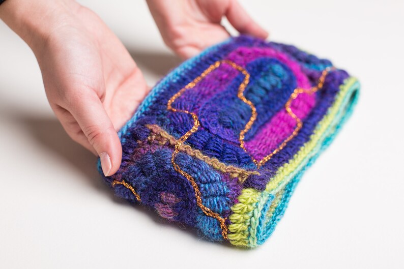May include: A colorful knitted headband with a blue, green, purple, and pink color scheme. The headband is decorated with gold thread.
